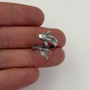 May include: A silver-colored ring in the shape of a fish. The ring wraps around the finger, with the fish's head at the top and the tail at the bottom. The fish has detailed scales and fins, and the ring appears to be made of a shiny metal.