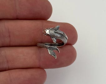 May include: A silver fish-shaped ring. The ring wraps around the finger, with the fish's head at the top and the tail at the bottom. The fish has detailed scales and fins, and the ring appears to be made of a shiny metal.