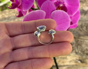 May include: Silver ring with three mushroom-shaped details. The ring is on a hand against a background of pink orchid flowers.
