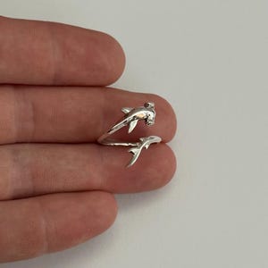 以下が含まれることがあります： ハンマーヘッドシャークのデザインが施されたシルバーカラーのリング。リングは指に巻き付き、サメの頭と尾が焦点となっています。リングは光沢のある金属素材で作られています。