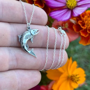 May include: A silver necklace with a detailed silver fish pendant. The necklace is on a hand with a blurred background of colorful flowers.