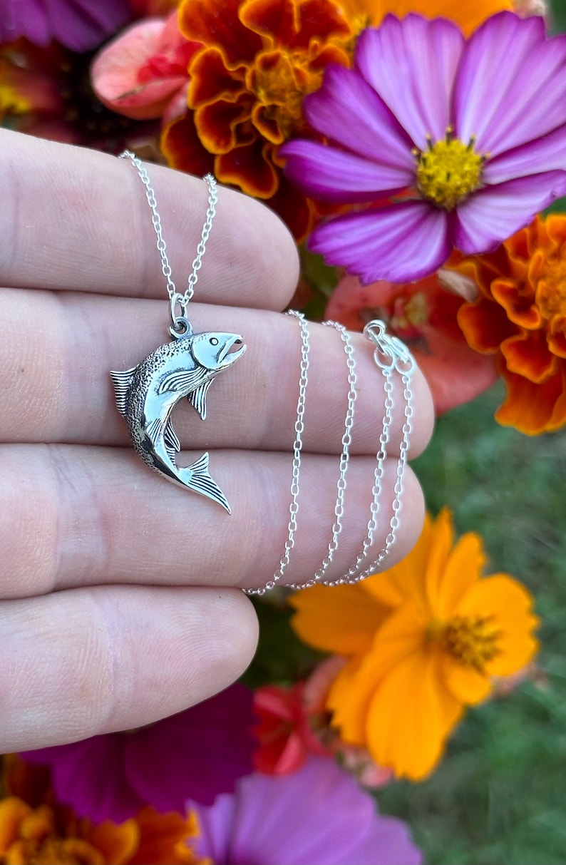 May include: A silver necklace with a detailed silver fish pendant. The necklace is on a hand with a blurred background of colorful flowers.