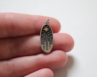 Puede incluir: Encanto ovalado de plata con una imagen en blanco y negro de un bosque con una estrella dorada en la parte superior.