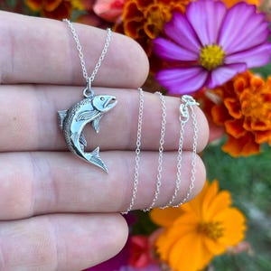 May include: A silver necklace with a detailed silver fish pendant. The necklace is on a hand against a background of colorful flowers.