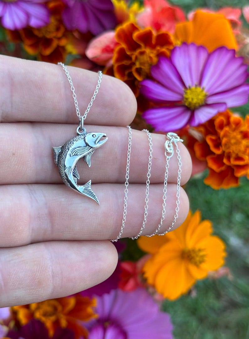 May include: A silver necklace with a detailed silver fish pendant. The necklace is on a hand against a background of colorful flowers.