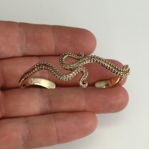 May include: A gold-colored, tentacle-shaped cuff bracelet. The bracelet has a detailed, textured surface with small, round suction cup details. The ends of the bracelet are smooth and open, designed to fit around the wrist. The bracelet is shown on a hand.