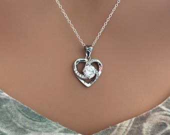 May include: A silver necklace featuring a heart-shaped pendant. The pendant has a central, round, clear gemstone surrounded by smaller, clear stones and a heart-shaped frame. The necklace chain is delicate and silver.