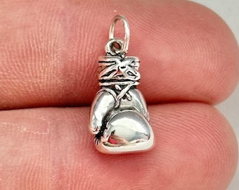 May include: Silver charm in the shape of a boxing glove. The glove is wrapped with tape and has a loop at the top for attaching to a bracelet or necklace.