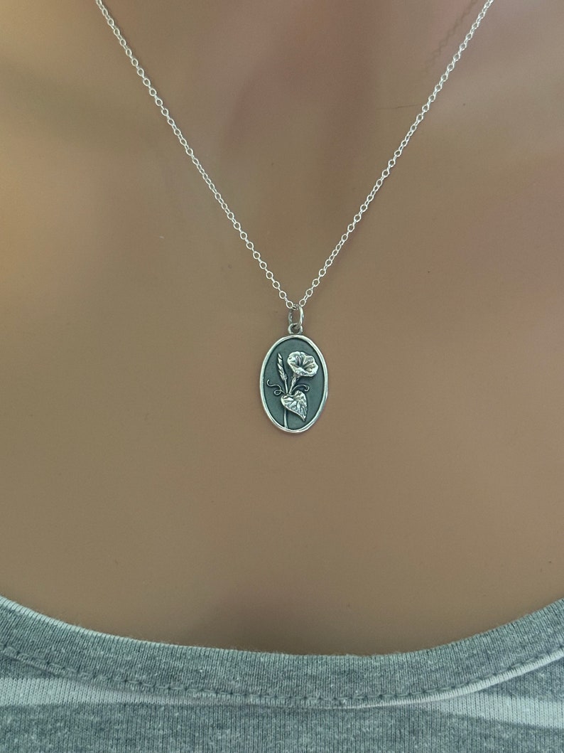 Peut inclure: Un collier en cha&icirc;ne argent avec un pendentif ovale en argent grav&eacute; d'un motif floral. La fleur est blanche avec une tige et des feuilles gris fonc&eacute;.