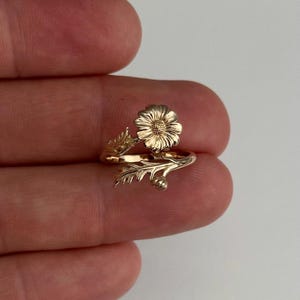 May include: A gold-colored ring featuring a detailed daisy flower design. The ring wraps around the finger with delicate leaf and bud accents. The floral theme is prominent, showcasing intricate craftsmanship.