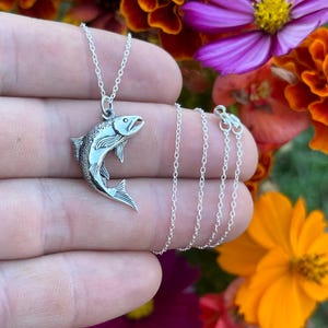 May include: A silver necklace with a detailed silver fish pendant. The necklace is on a hand against a background of colorful flowers.
