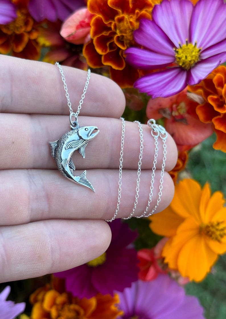 May include: A silver necklace with a detailed silver fish pendant. The necklace is on a hand against a background of colorful flowers.