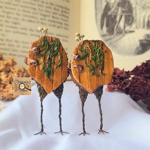 May include: Two whimsical, handcrafted wooden sculptures. Each features a shield-shaped wooden body with green moss and small snail details. The sculptures stand on delicate wire legs, evoking a fantasy theme.