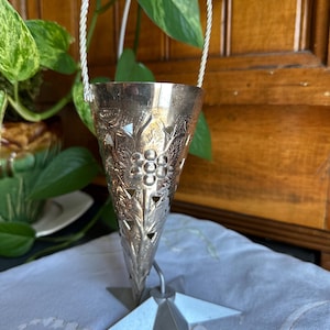 May include: A silver, cone-shaped tea infuser with a star-shaped base and a twisted rope handle. The infuser has a decorative design of leaves and grapes.