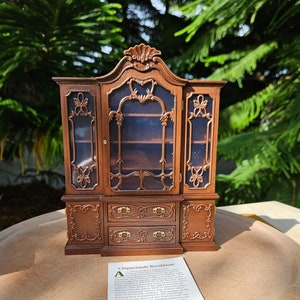 May include: A miniature wooden Chippendale breakfront with glass doors and drawers. The breakfront is made of dark wood with intricate carvings and gold accents. The text on the card reads: "A distinctive Chippendale Breakfront which is a great challenge to build. The breakfront is made of mahogany with a walnut inside and two great display areas. The doors are fitted with glass and the drawers are fitted with brass pulls. This edition. The doors are held in place by a unique system of hinges. The drawers are fitted with brass pulls. The intricate scrolling designs, the meticulous hand-cut details, and the fine craftsmanship make this breakfront a true collector's item. The breakfront is 12 inches high, 8 inches wide, and 4 inches deep. The breakfront is a great addition to any dollhouse or miniature collection."