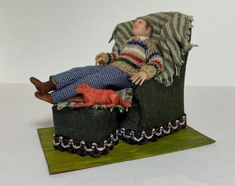 May include: A miniature dollhouse scene featuring a person sitting in a green armchair with a red dog on a footstool. The armchair has a striped throw blanket and the footstool has a patterned fabric.