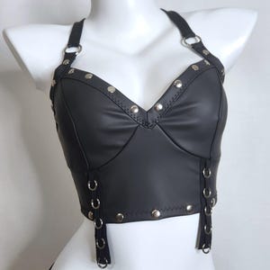 May include: Black faux leather halter top with silver-tone metal studs and grommet details. The top features a V-neckline and adjustable straps. The garment is displayed on a white mannequin.