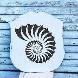 May include: A black and white nautilus shell stencil on a white wooden background. The stencil is shaped like a spiral.