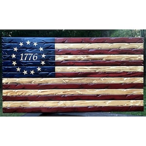 May include: A wooden American flag with 13 red and white stripes and a blue canton with 13 white stars in a circle. The canton also has the year "1776" inscribed in gold lettering.