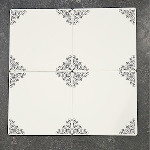 May include: Four off-white ceramic tiles arranged in a square, each with a gray floral corner design. The center of the square features a larger, ornate gray floral pattern. The tiles are set against a dark gray surface.
