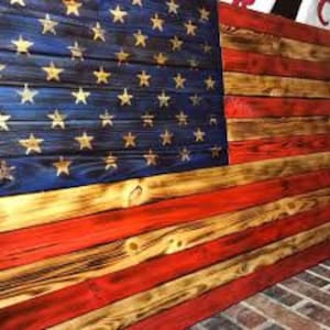 May include: A wooden American flag with a distressed, weathered look. The stars are gold and the stripes are red, white, and blue. The flag is made of reclaimed wood and has a rustic, vintage feel.
