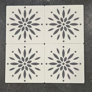 May include: Four square, off-white ceramic coasters with a gray floral design. Each coaster features a central flower-like pattern with radiating petal shapes and small dots at the corners. The coasters are arranged on a dark, textured surface.