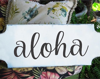 May include: White wooden sign with a distressed finish and rounded edges. The sign has the word "aloha" written in black cursive lettering.