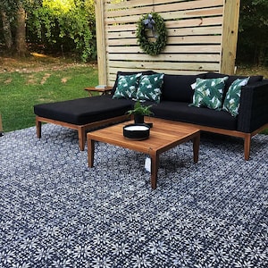 May include: A black sectional sofa with green leaf patterned throw pillows sits on a patio with a black and white patterned concrete floor. A wooden coffee table sits in front of the sofa with a black candle holder on top. A wooden bench with a black cushion sits to the left of the sofa.