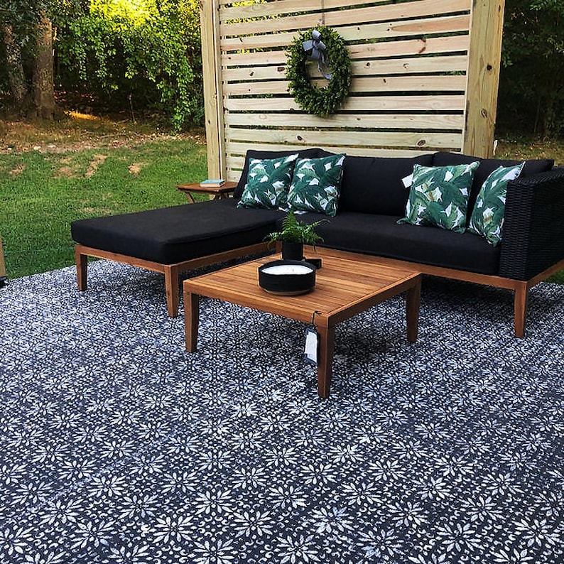 May include: A black sectional sofa with green leaf patterned throw pillows sits on a patio with a black and white patterned concrete floor. A wooden coffee table sits in front of the sofa with a black candle holder on top. A wooden bench with a black cushion sits to the left of the sofa.