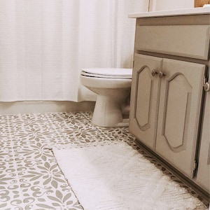 May include: A white toilet with a closed lid sits in a bathroom with a white shower curtain and a gray cabinet with two doors. The floor is tiled with a brown and white patterned tile.