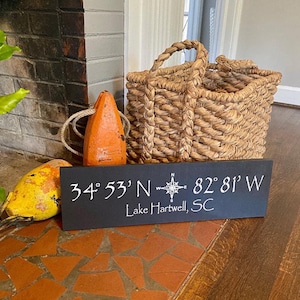 May include: Black wooden sign with white text that reads "34° 53' N - 82° 81' W Lake Hartwell, SC". A compass rose is between the coordinates. The sign is on a tiled surface with a woven basket and orange buoy.