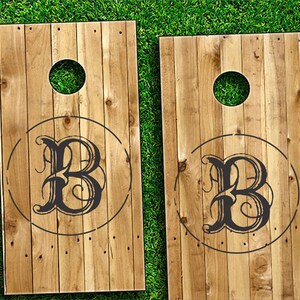 Op de afbeelding: Twee houten cornhole-borden met een zwart-wit monogram van de letter "B" in een cirkel op elk bord.