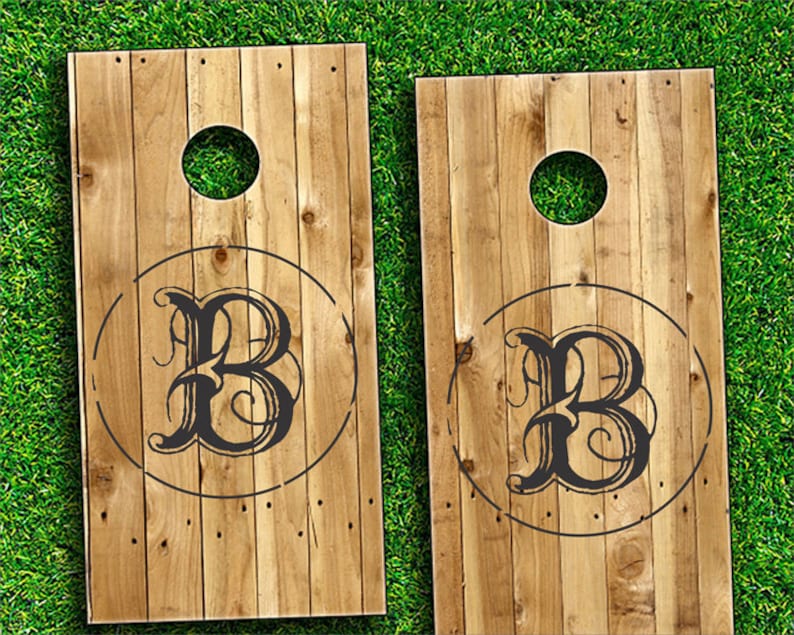 Op de afbeelding: Twee houten cornhole-borden met een zwart-wit monogram van de letter "B" in een cirkel op elk bord.