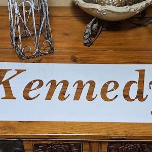 May include: A white stencil with the name "Kennedy" cut out in a serif font. The stencil is on a wooden surface. The dimensions are 44" x 22.5".