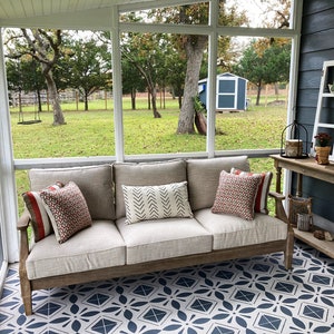 May include: A light beige sofa with pillows in red, white, and black patterns sits on a screened porch with a patterned tile floor. The porch has a view of a green lawn and trees.