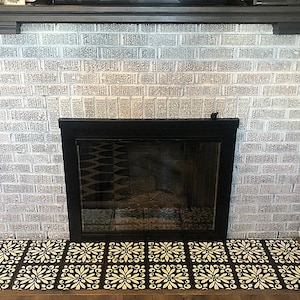 May include: A white brick fireplace with a black metal fireplace screen and a decorative tile hearth. The tiles are black with a white floral pattern.