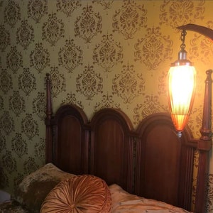 May include: A wooden bed frame with a brown headboard and a gold-colored lamp with a glass shade hanging from the ceiling. The wall behind the bed is covered in a yellow and brown damask patterned wallpaper.