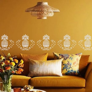 May include: A living room with a mustard yellow wall and a brown couch. The wall has a white stencil design of pineapples along the bottom. The couch has two pillows, one is a solid gold color and the other has a floral pattern. There is a coffee table in front of the couch with a vase of flowers and a few decorative items.