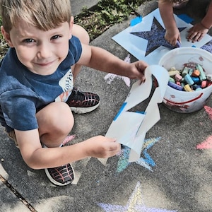 May include: A young child kneels on a concrete surface, holding a stencil over a colorful chalk star drawing. A bucket of chalk and another child are in the background. The child is wearing a blue shirt and black and red sneakers.