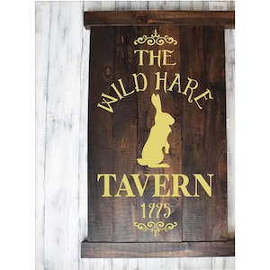 May include: A rustic wooden sign with the text "The Wild Hare Tavern 1775" in gold lettering. A silhouette of a yellow hare is in the center of the sign.