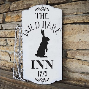 May include: A white wooden sign with black text and a silhouette of a rabbit. The sign reads "THE WILD HARE INN 1775" with decorative flourishes. The sign is mounted on a decorative metal frame.