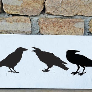 May include: A rectangular white wooden sign featuring three black crow silhouettes. The crows are in various poses, with one appearing to be cawing. The sign is set against a rustic brick background.