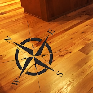 May include: A black compass stencil on a wooden floor with the letters N, S, E, and W.