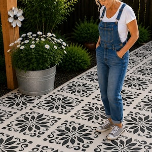 May include: A white t-shirt and blue denim overalls paired with gray canvas sneakers. The outfit is worn while standing on a light-colored patio surface featuring a repeating black floral stencil pattern, with a metal planter nearby.