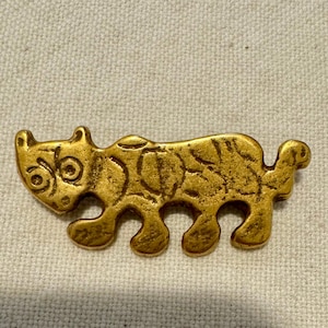 Puede incluir: Figurilla de tigre estilizada dorada con superficie texturizada. El tigre tiene rasgos redondeados, incluyendo una cabeza con ojos circulares y cuatro patas. La figurilla es probablemente de metal.