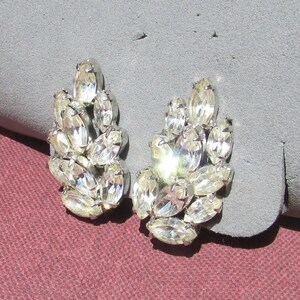 May include: A pair of silver-toned clip-on earrings. Each earring features a cluster of clear, oval-shaped rhinestones. The rhinestones are arranged to create a floral or leaf-like design, reflecting light and adding sparkle. The earrings are set against a gray background.