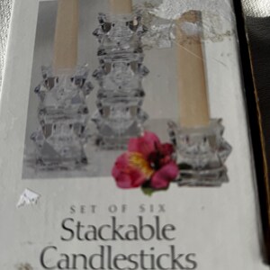 May include: A box set of six stackable lead crystal candlesticks. The clear glass candlesticks have a starburst design. The box has the word "Tuscany" and the words "Set of Six Stackable Candlesticks" printed on the front.