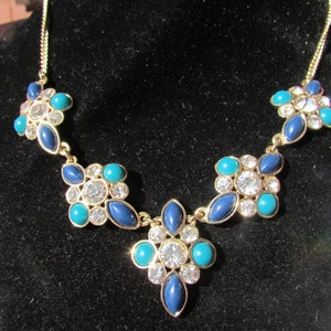 May include: A gold-toned necklace with a pendant design. The necklace features clusters of blue, turquoise, and clear crystal-like stones. The necklace is displayed on a black velvet surface.