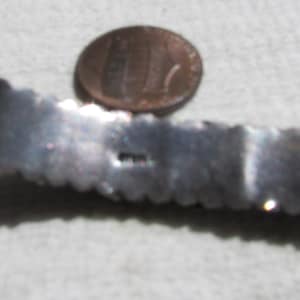 May include: Close-up of the back of a silver bracelet with a textured design. The bracelet is about the size of a US penny.