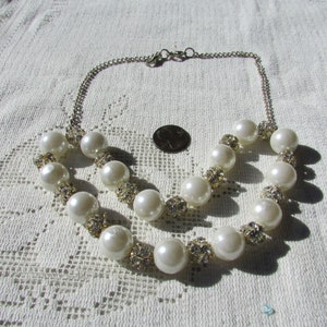 May include: A double-strand pearl necklace with large, round, off-white faux pearls. The necklace features silver-toned chain and accents of small, sparkling rhinestones. The necklace is displayed on a white, lace-patterned surface.